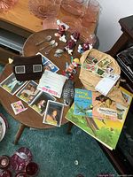 Overview photo of collectibles on a small round wooden table including grapevine figures, View-Master, trading cards, sticker books, and pink glass dishware in the background.