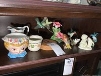 Photo of assorted collectibles including Dept 56 Snowbaby planter, small pitcher, and bird figurines displayed on dark shelf