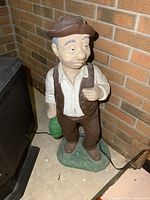 Photo of full concrete statue of man wearing a brown vest, pants, white shirt, and brown hat, holding a green object, placed near a brick wall indoors.
