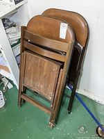 Photo showing a group of three folding chairs leaning against a wall including two metal folding card table chairs with brown vinyl padded seats and backs and one wooden folding chair with a slatted seat and backrest.