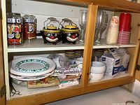 Wide shot of contents inside cupboard showing vintage clown sugar and cream containers, disposable plates, Sunday Funnies glasses, multiple wine glasses, and Ekco hand mixer box.