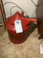 Front side view of the red painted metal kerosene can showing the spout, handle, and overall shape with inventory tag.