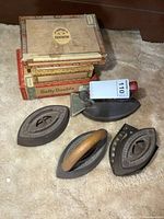 Four antique irons with various designs and one cast trivet, together with a stack of vintage cigar boxes. Irons show rust and age.