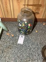 Full view of large clear glass jar filled with assorted vintage marbles placed on carpeted floor near wood paneling.