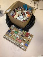 Full view of large cardboard box filled with loose matchbooks and a wooden display board with mounted matchbooks beneath the box.