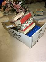 Large white cardboard box filled with multiple decorative smaller boxes containing matchbooks and matchbook covers, placed on a floor with some floral arrangements in the background