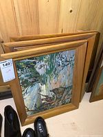 Three oil on board paintings stacked and leaning, wood frames visible, nature scene with man and dog shown