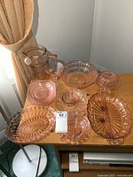Photo showing a collection of pink depression glass items including pitcher, plates, bowls, butter dish, and serving dishes on table