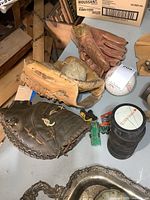 Photo showing three vintage leather baseball gloves with signs of wear, two baseballs, four black hockey pucks stacked, and some die-cast cars in the background.