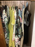 Multiple vintage ladies square dance dresses and skirts hanging on wooden hangers in a closet. Various colors and fabrics including floral, black with silver, yellow, and multicolored prints.