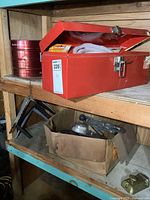 Photo of red metal toolbox on wooden shelf with emergency kit items inside. Cardboard box with tools below.