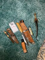 Top view photo of all items including two cutters with sheaths, souvenir folding pocket knife, miniature dagger with decorated sheath, and wooden pocket tool lined on carpet.