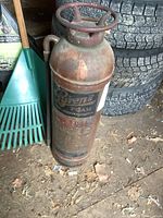 Full view of vintage Pyrene copper fire extinguisher standing upright on ground next to green rake and tires