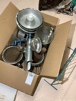 Box containing assorted vintage kitchenware including a metal food mill, sieve, and wooden-handled potato masher inside a cardboard box.