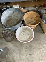 Wide view showing large corrugated metal washtub, wooden apple basket, white enamel pan, and cast iron kettle on floor.