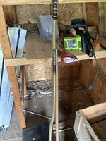 Wide shot showing Poulan electric chainsaw on wooden shelf with shovel standing upright in front and leaf rakes on floor.