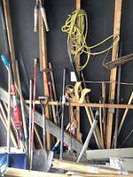 Sun Joe electric pole chain saw, shovels, wooden handles, rope, axe, and other tools hanging on a wall.