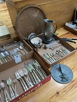 Photo showing overall assortment of flatware in boxes, hammered pitcher, metal tray, turkey figurine and sundial on wooden table