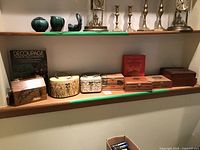 Wide view of shelf displaying eight decorative boxes and two books, predominantly made of wood, metal, and decoupage materials.