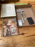 Overview of all items including Holy Bible, New Testament Story book, crucifix, medal, and small black book placed inside a cardboard box