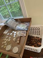 Mixed coins, French banknotes, silver coins in plastic cases, laid out in a cardboard box and white basket on the floor near a window.