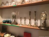 Display shelf with two anniversary clocks under glass domes, four brass candlesticks, two small green Blue Mountain pottery pieces, and decorated porcelain items on top shelf