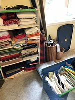 Overview of white shelving unit stacked with folded fabrics in various colors and patterns, plastic and wicker containers nearby with additional fabrics.