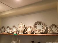 Full view of the entire cocoa set displayed on a shelf showing 6 cups with saucers, 6 plates, coffee pot, sugar bowl, and creamer with floral pattern and gold accents.