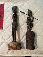 Photo of two wooden warrior sculptures on a mattress surface, showing full height and detail.