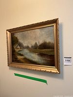 Full view of the oil painting 'House Next to a River' by J. Burton in an ornate gold frame mounted on a wall.