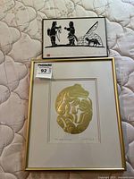 Two framed Indigenous prints shown together on a mattress, one black and white by Henry Napartuk and one gold embossed by Bill Reid