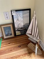 Photo showing both framed sailing art prints and two wooden model boats together on hardwood floor against wall corner.