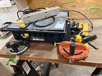 Front side view of Mastercraft wet tile saw showing ribbed silver cutting platform with power cord and yellow adjustment knobs, includes orange and black angle guide attachments with handles and degree markings.