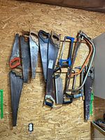 Entire lot showing 10 saws lying on a wooden surface with visible wear and rust. Includes handsaws with wooden and plastic handles, hacksaws with metal frames, and crosscut saws with large blades.
