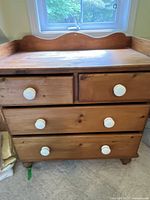 Front view showing the full dresser with four drawers and white ceramic handles. The dresser has a scalloped backboard and visible surface wear.