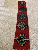 Full length view of narrow Navajo rug runner on carpeted floor showing red background with geometric shapes in teal, yellow, and black.