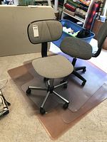 Two secretarial office chairs, one light grey with rectangular backrest and one darker grey with rounded backrest, both on caster wheels, placed on clear plastic under desk mats.