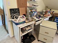 Two-drawer filing cabinet positioned beside a desk loaded with various office supplies on top and in drawers and shelves.