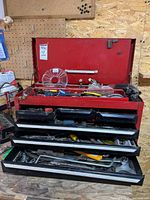 Open red metal toolbox showing contents including multiple tools in three black drawers and top compartment.
