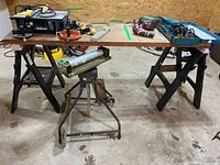 Wide view of two sawhorses holding a wooden tabletop with various tools on top, and an adjustable height roller support beneath the tabletop.