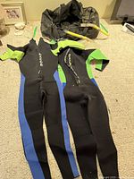 Two wetsuits made by Dacor, black with green and blue panels, front zipper closure item on floor, with snorkels and a mesh bag behind