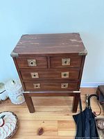 Front view of a small wooden chest of drawers showing metal ring pull handles and metal corner accents on the top corners. Surface wear is visible on the top wood surface.