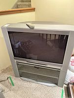 Front view of Toshiba 34 inch CRT TV showing the glass screen and built-in storage below the screen with glass doors. Remote control placed on top.