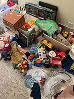 Wide view photo showing collection of vintage toys including Fisher Price garage, various dolls, books packed in bins, and toy train accessories scattered on carpeted floor.
