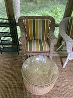 Wicker armchair with striped cushion and round wicker table with glass top in front on a natural fiber rug.
