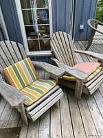 Two weathered wood Adirondack chairs with striped green, orange, yellow, and white cushions. Placed on wooden deck in front of windowed house door.