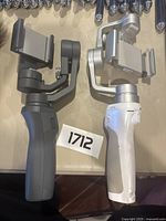 Two DJI Osmo handheld gimbals laid side by side on a flat surface, one grey, one white with silver clamps.