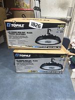 Two stacked boxes of Topaz LED round high bay black power selectable lights showing front packaging indicating product features and model.