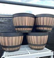 Four large plastic barrel-style planters with wood grain pattern and black rims and bands, displayed outdoors on a white support.