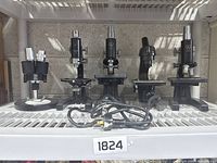 Front view of the five microscopes arranged on metal shelves, showing various sizes and styles, all black metal with adjustable parts and cords.